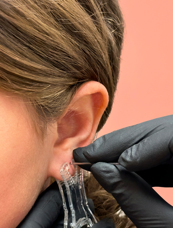 Person getting an earring put into their ear with a pink background